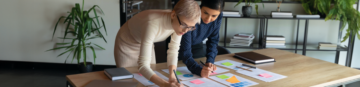 Two people collaborate in an office, reviewing colorful charts on a wooden table. Books and plants decorate the modern work environment.