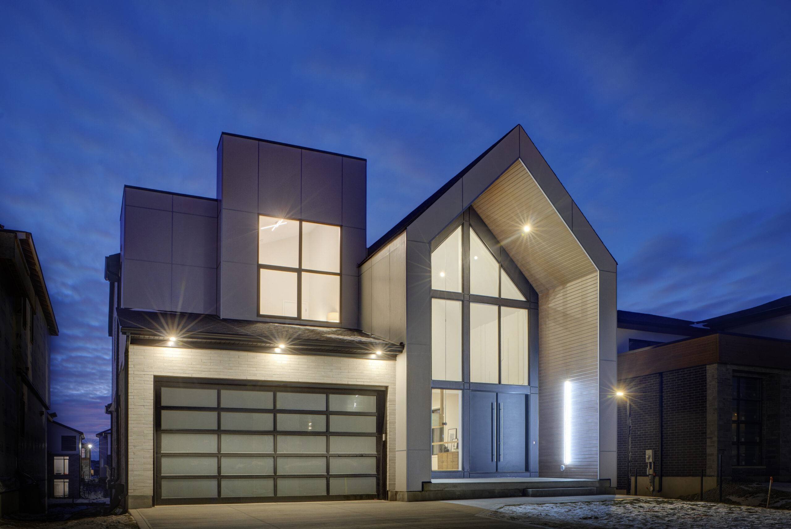 A modern two-story house with a white facade, a pitched roof with a glass window, and a garage door with glass panels. The house is illuminated at night, and the sky is dark blue.