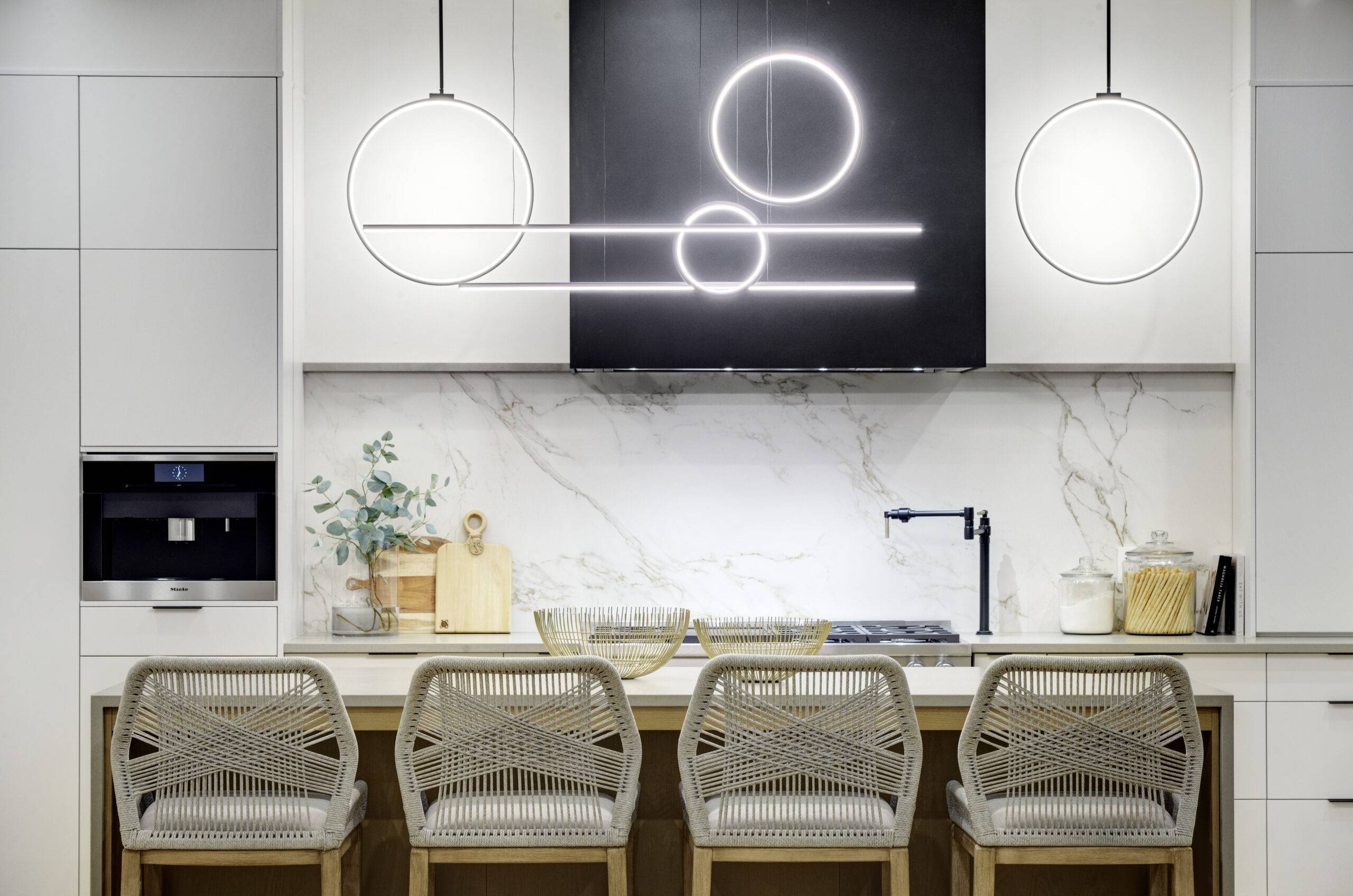 A modern kitchen with white cabinets, a white countertop island, and elegant flooring. Four woven bar stools sit at the island, and a black hood with geometric lighting hangs above the stove.