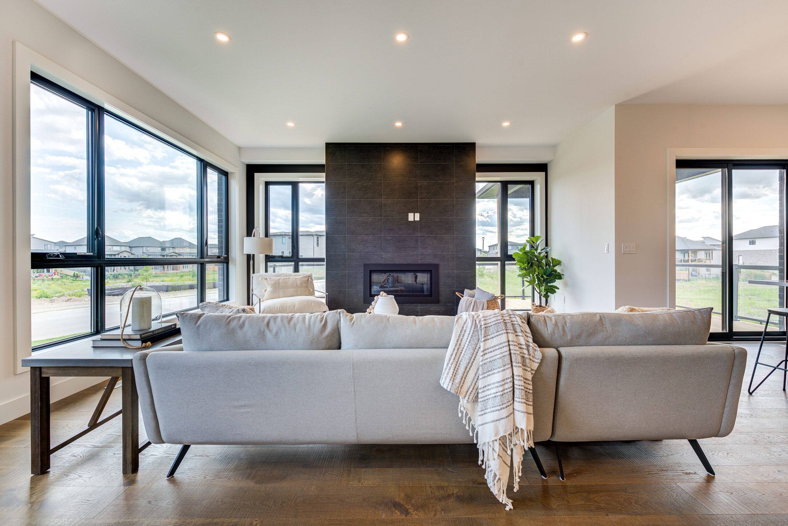 A modern living room with a gray fireplace, large windows, and elegant flooring. A white sofa sits in front of the fireplace with a throw blanket draped over it.