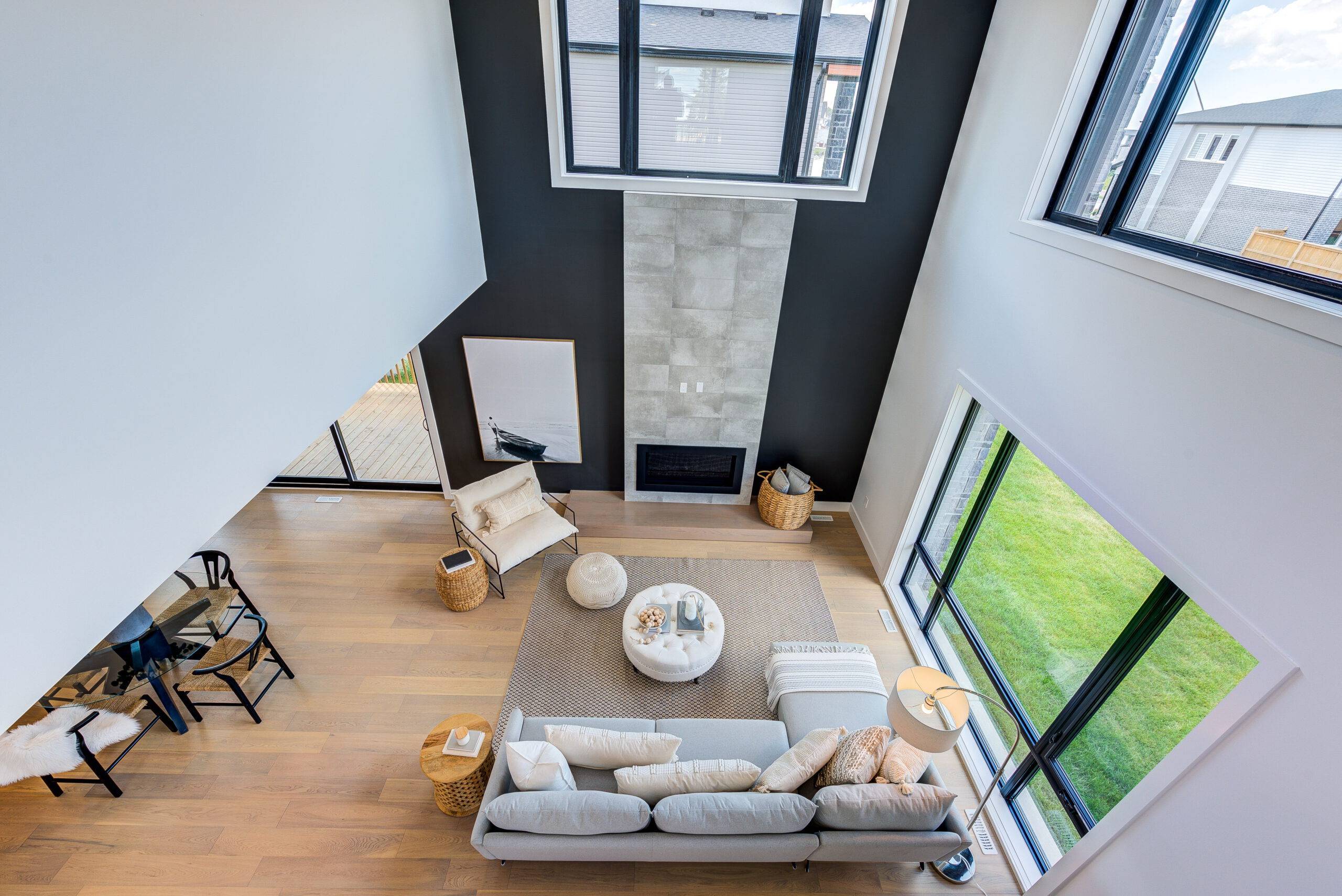 A spacious two-story living room with a white sofa, a fireplace, and large windows. A chair and ottoman sit by the fireplace, and a dining table is visible in the background.