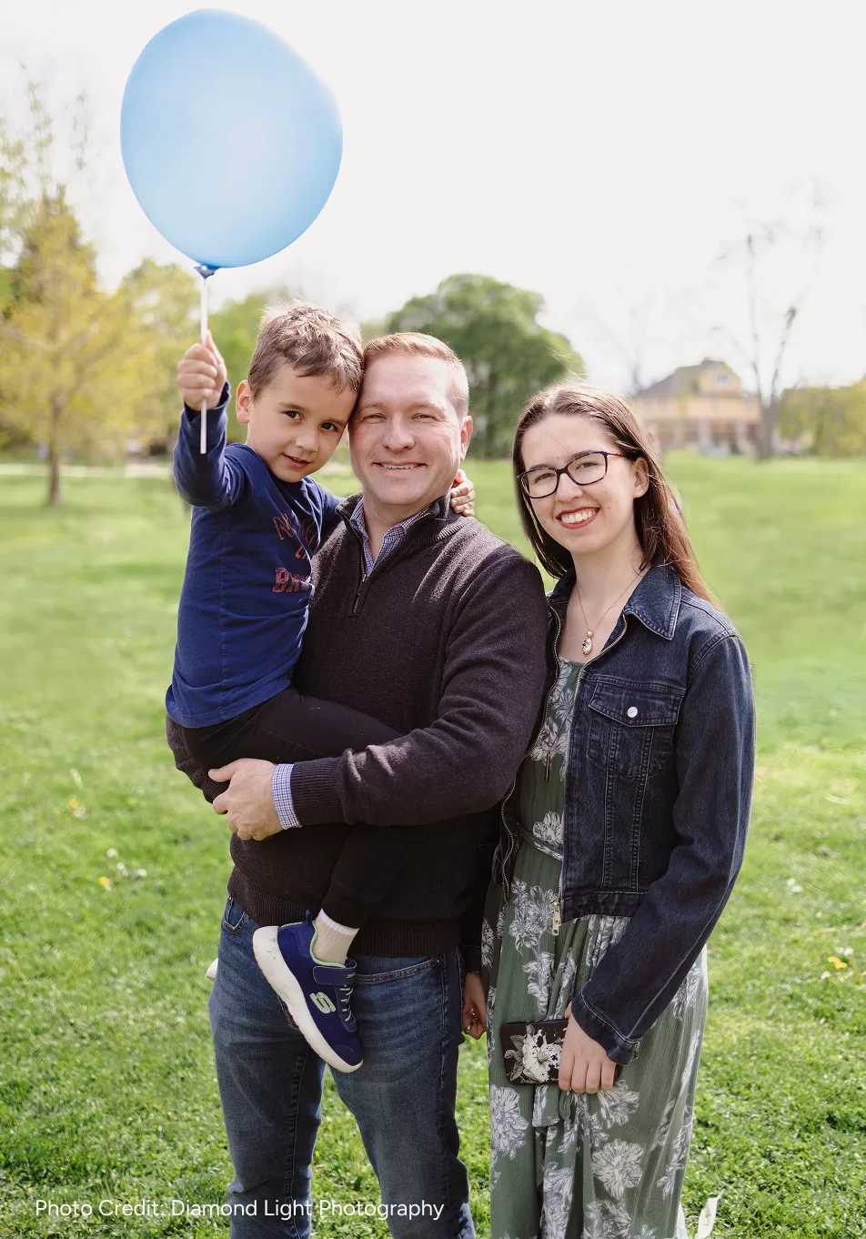 Josh Morgan and two of his kids standing on a green lawn. He is holding his son with a blue balloon, while his smiling daughter stands beside them. The background is a bright park. Photo Credit: Diamond Light Photography
