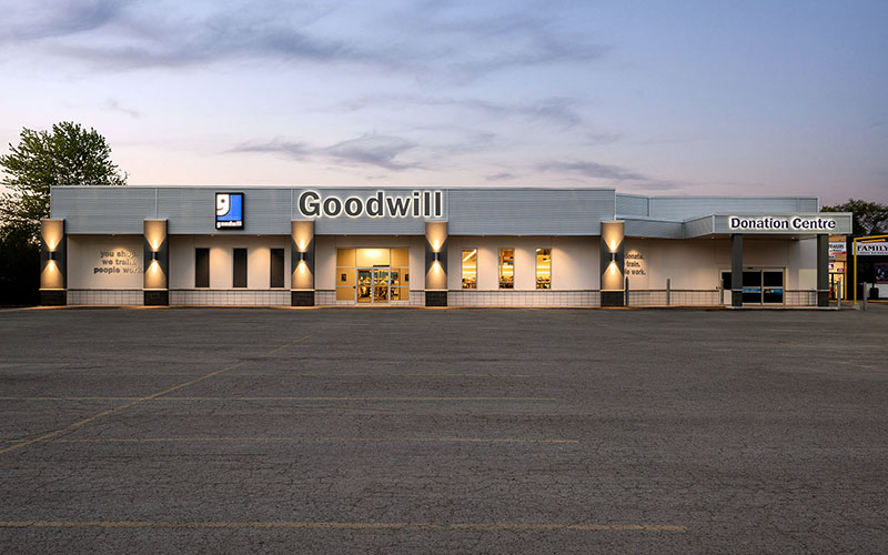 Commercial construction project, the front of Goodwill Store, London, Ontario showing the whole building, 5 grey columns spaced along the front with lights turned on, the Goodwill sign above the front in the center on a light blue background with the logo beside it.