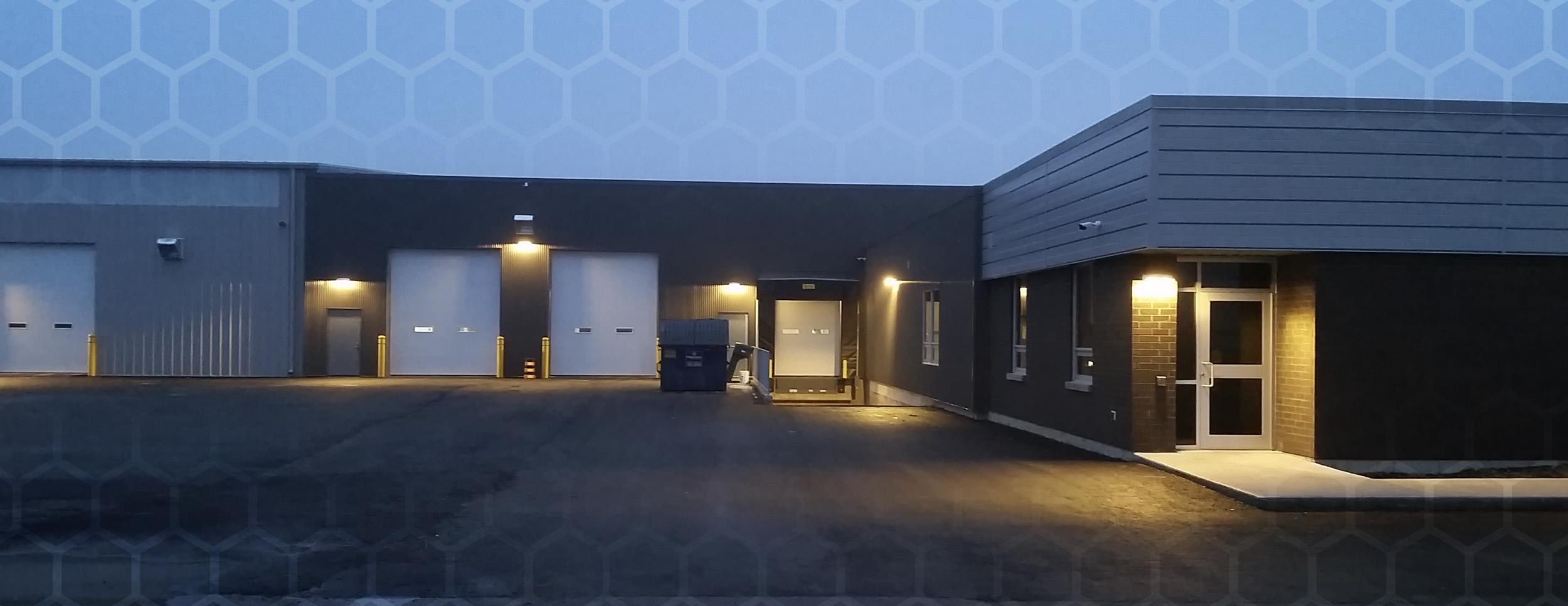 Evening Photo of the exterior of the Weathertech Building, sides are grey brick with grey metal cladding around the top with 4 white garage doors for loading