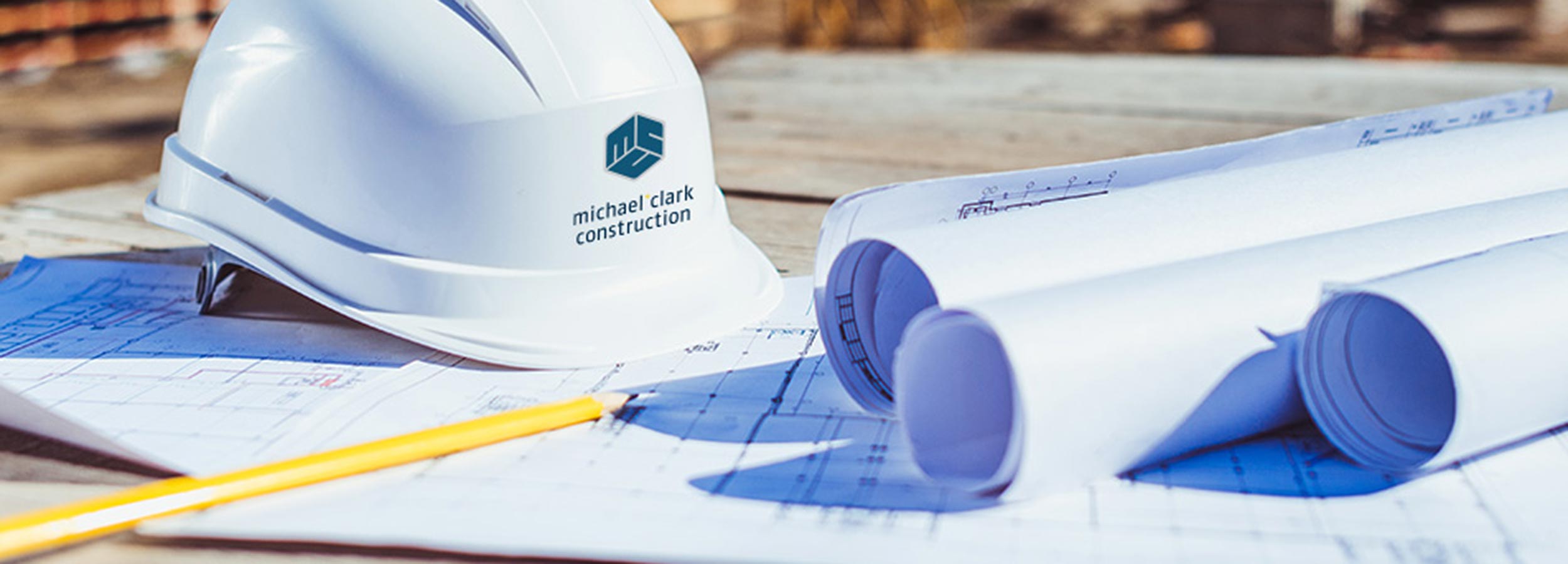 Picture of a white hardhard with company logo, sitting on a blueprint with other blueprints rolled up.