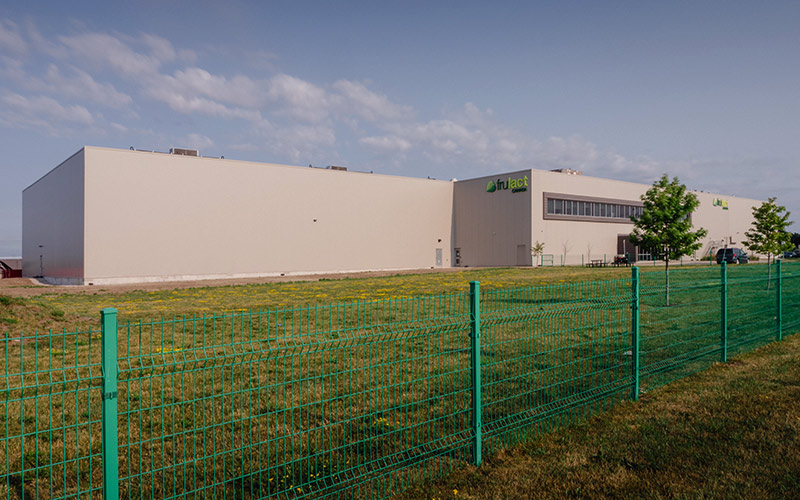 exterior view of Frulact building, beige metal siding and green metal fence surrounding property.