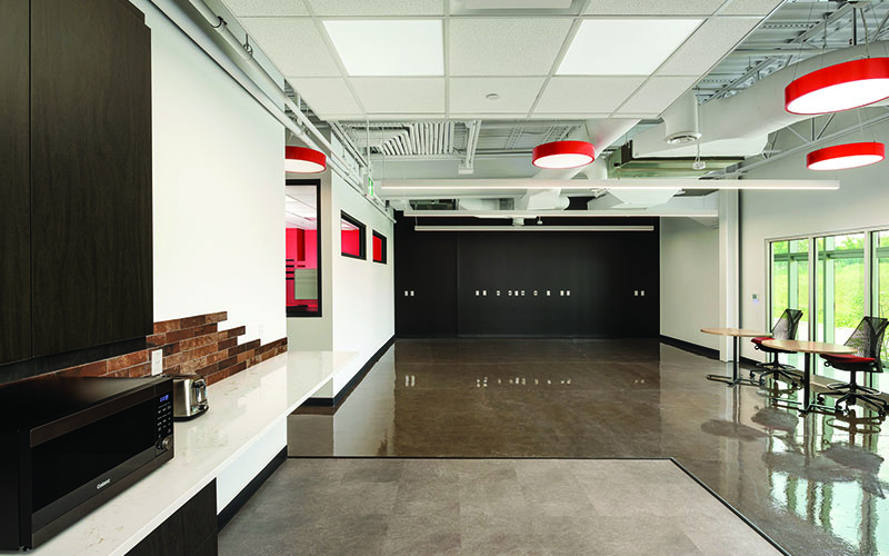 BOS Innovations at The Collider Western University Research Park, Photo of an interior space, open concept lunhc room, 2 white walls, large circular red pendelum lights hanging from the ceiling, glass windows on the left wall with chairs in front.