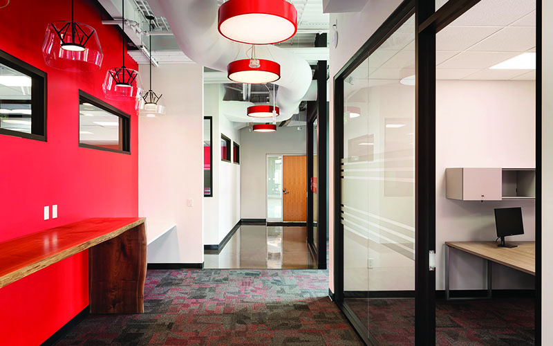 BOS Innovations at The Collider Western University Research Park, Photo of an interior hallway, Red wall on left side with partial long rectangular window three quarters of the way up, large red circular lights hanging from the ceiling in a row down the center of the hallway, floor to ceiling glass walls on the right