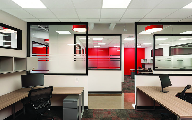 BOS Innovations at The Collider Western University Research Park, Photo of an interior space, showing the back of two desks, entrance in to the hallway in the middle and glass wall on the other side with red walls in the distance.