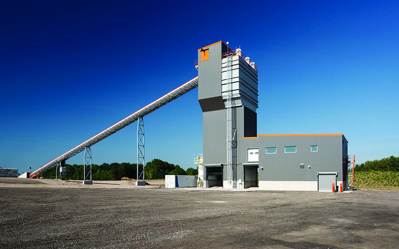 Photo of a concrete plant industrial facility, blue skies and it is in the middle of a open space. Grey L-shaped building standing towards the sky.