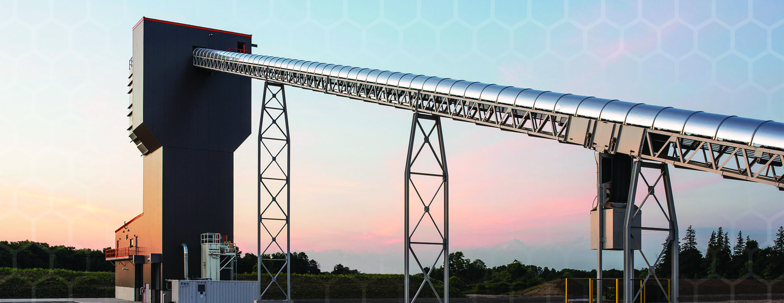 Evening Sunset Photo with a large steel round conveyor raising from the ground on a slant to the top of a tall building. The conveyor is supported by 2 steel tall supports at 2 intervels as it inclines.