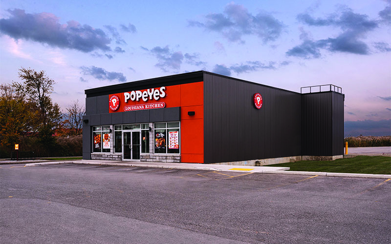 Side Rendering of Exterior Rendering of a Popeyes Restaurant at Sunset. Dark Grey brick with pops of Red along the top and down the right side of the front of the building. Dark grey for the side wall of the building.