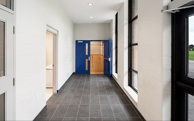 Covenant School, interior photo of a hallway leading to the gym in the distance with white cinder block walls and grey tiled flooring. Royal Blue doors flank the entrance to the gym.