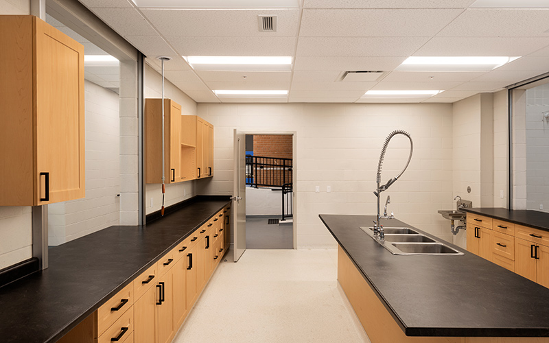 Covenant School, interior photo of kitchen with light oak cabines along the left and right, black laminate countertop with black drawer handles and black door handles. In the middle of a room is an island of light oak with black laminate counter top, triple sink in the center and a hand held extendible faucet.