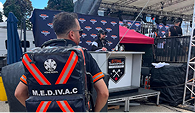 A person wearing a black and orange MEDIVAC backpack stands on a paved area near a stage with a "Stihl Timbersports" banner. The scene has an outdoor event atmosphere.