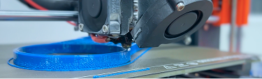 A 3D printer nozzle is depositing blue filament, creating a circular object on the printer bed.