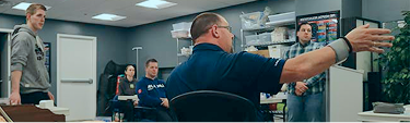 A man seated at a desk gestures while speaking to four people standing and sitting in an office with shelves and supplies in the background.