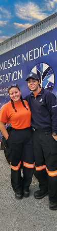 Two Mosaic Medical medics in orange and navy uniforms stand smiling in front of a medical station sign under a blue sky, conveying teamwork and professionalism.