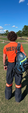 A Mosaic Medical medic stands in a grassy field under a blue sky, wearing an orange "MEDIC" shirt and carrying a blue backpack, conveying readiness and alertness.