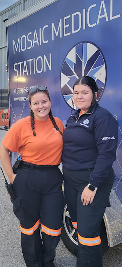 Two women in medical uniforms stand smiling in front of a mobile medical station. The backdrop has "Mosaic Medical Station" and a medical logo.
