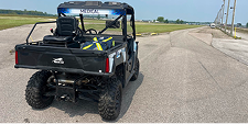 A black utility vehicle with a flashing light bar on a rural road. The road is clear and stretches into the distance under a clear blue sky.