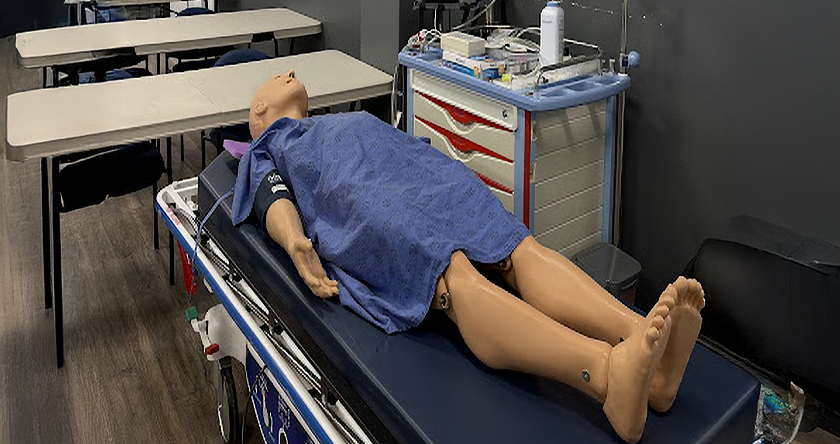 A medical training room with a human-like mannequin on a stretcher, draped in a blue hospital gown. Nearby are tables, chairs, and medical equipment.