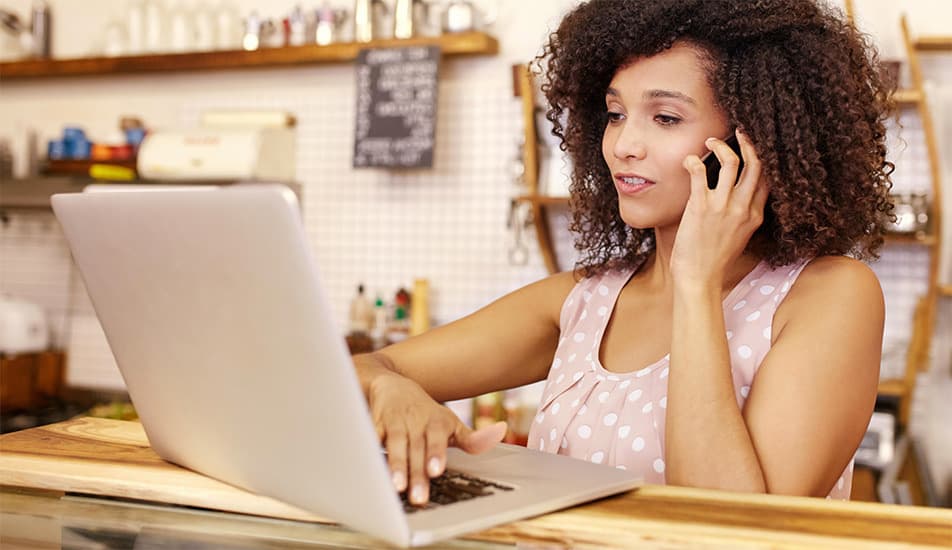 A woman with curly hair, wearing a pink polka-dot top, talks on the phone while typing on a laptop in a cozy café, conveying focus and multitasking.