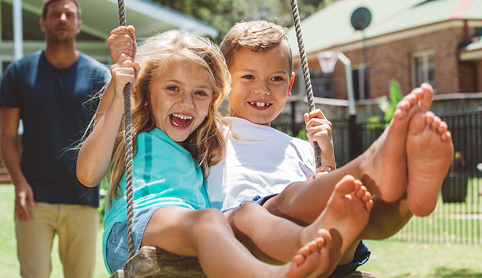 Two joyful children, a girl and a boy, swing together in a sunny backyard, smiling brightly. An adult stands behind them. The atmosphere is playful and carefree.