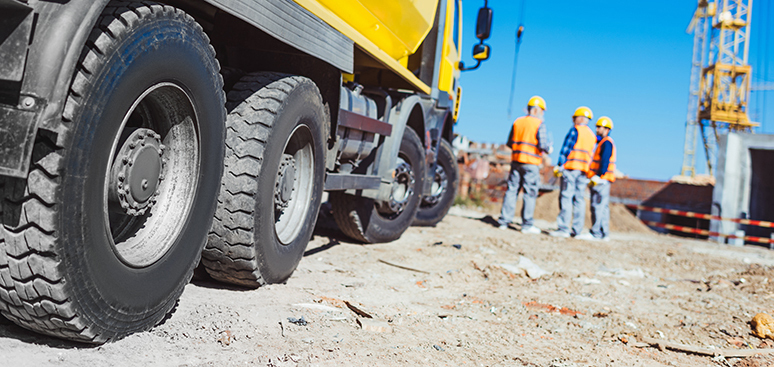 Close-up of large truck tires on a construction site, with three workers in helmets and safety vests discussing plans in the background. Clear blue sky.