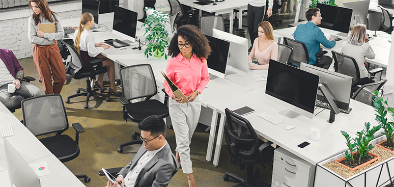 A busy open-plan office features diverse workers at computers, engaged in discussions. A woman in a pink shirt walks purposefully, carrying a folder.