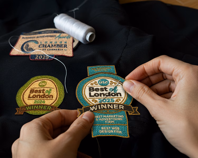 Hands sewing "Best of London" award patches onto fabric. A spool of white thread lies nearby. The scene conveys craftsmanship and achievement.