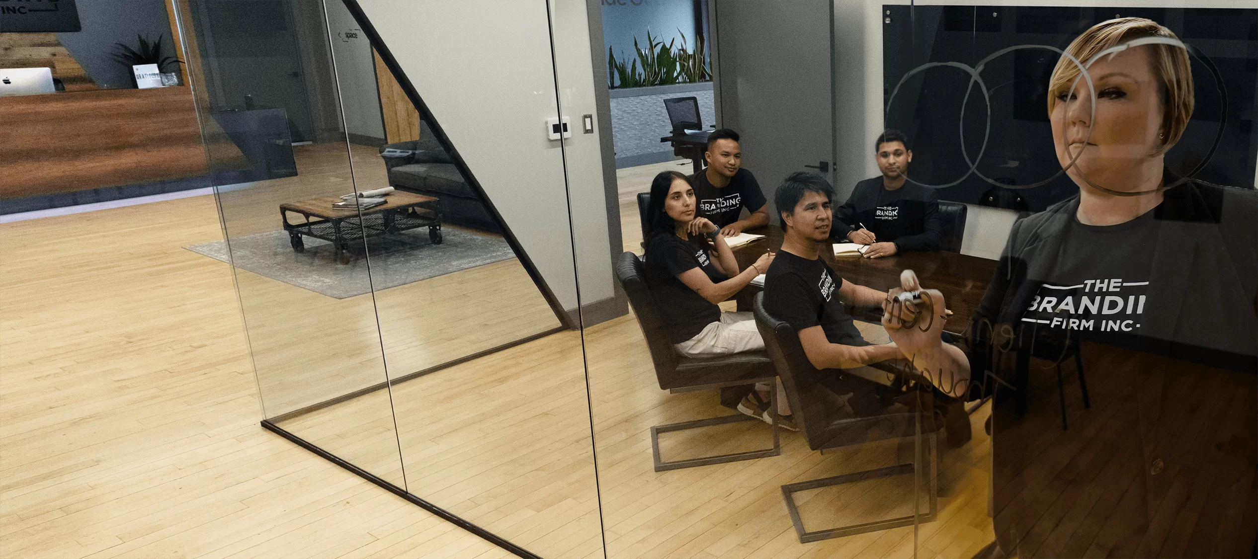 A group of four people sit at a conference table in a modern office with glass walls. They appear attentive, facing a speaker in a professional setting.