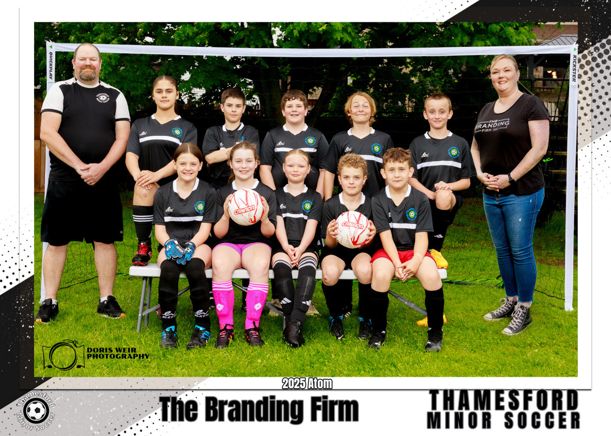 A youth soccer team poses on a field with two coaches. Players wear black jerseys and shorts, some with pink socks, holding soccer balls, smiling.